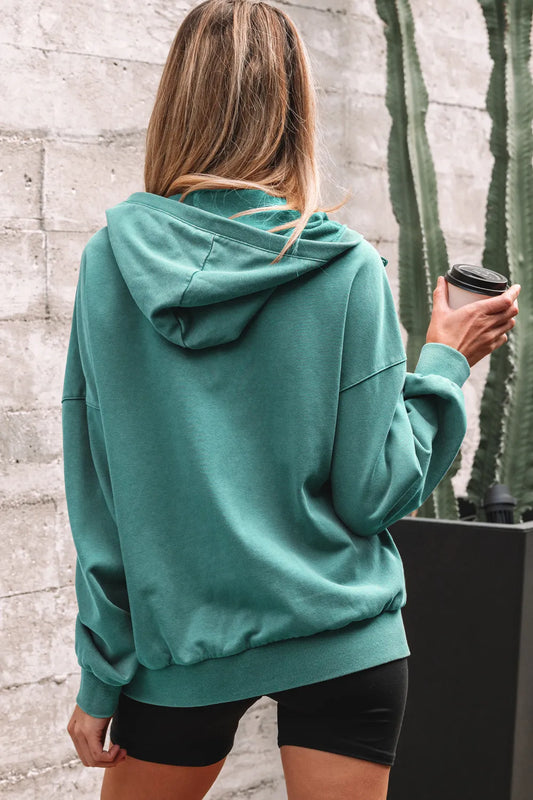 Person wearing a teal hoodie holding a coffee cup against a stone wall with a cactus plant.