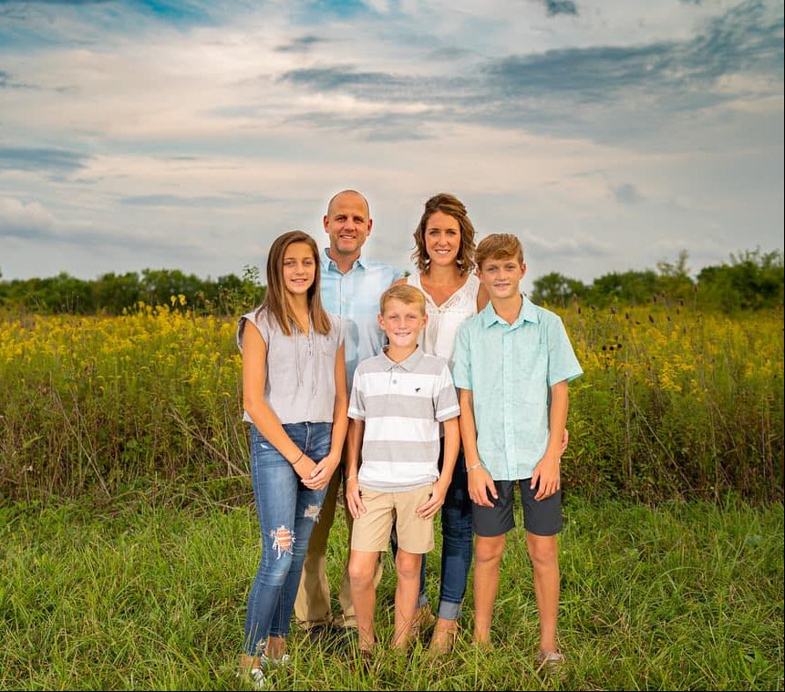 Family of five standing in a grassy field with a blue sky and clouds in the background.